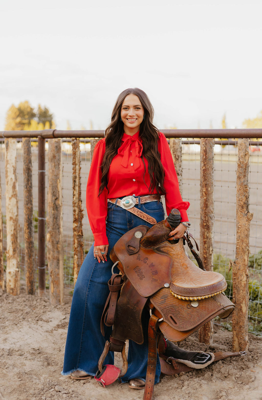 Woman in red blouse holding a western saddle by a fence.