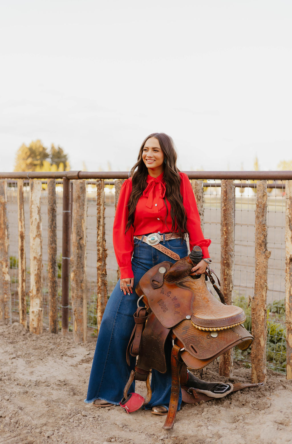 Woman in red blouse smiling and holding a saddle by a fence.