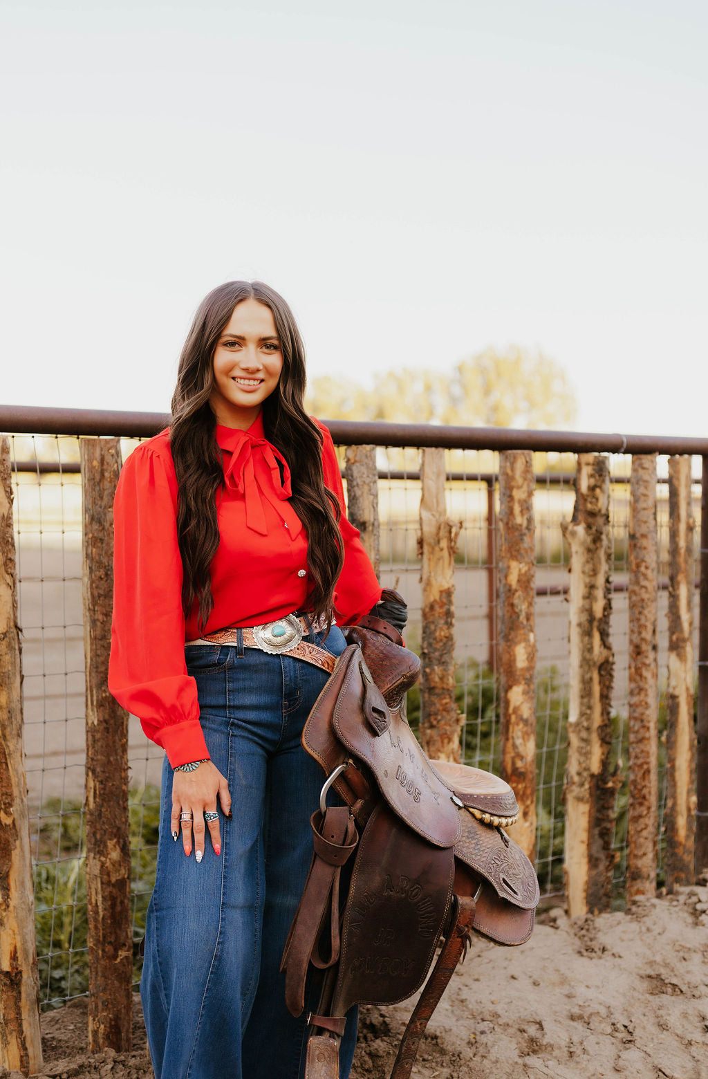 Woman in red blouse and flared jeans smiling while holding a saddle.