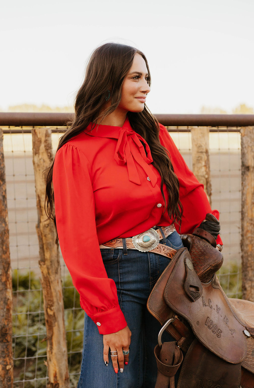 Woman in red blouse and jeans holding a western saddle, facing sideways.
