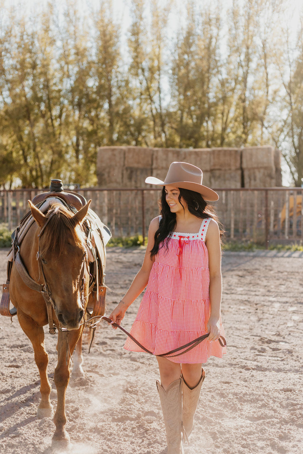 A horse and a woman holding its saddle lace, she's wearing the Sweet Land of Liberty Gingham Dress while they are walking