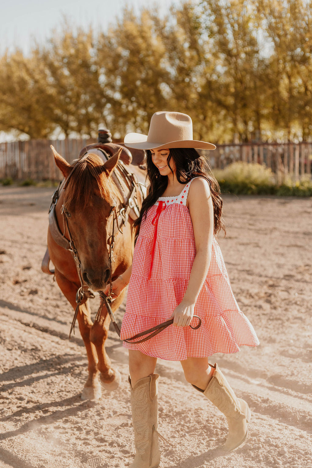 A horse and a woman holding its saddle lace, she's wearing the Sweet Land of Liberty Gingham Dress while they are walking